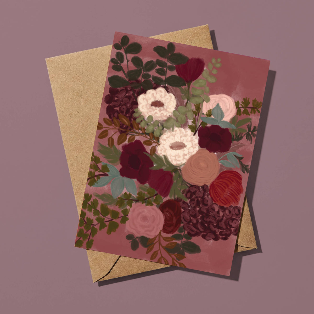 A floral art print card with a rustic kraft envelope, displayed on a solid mauve background. The card features a detailed botanical illustration of flowers in shades of burgundy, pink, cream, and green on a dusty rose background.