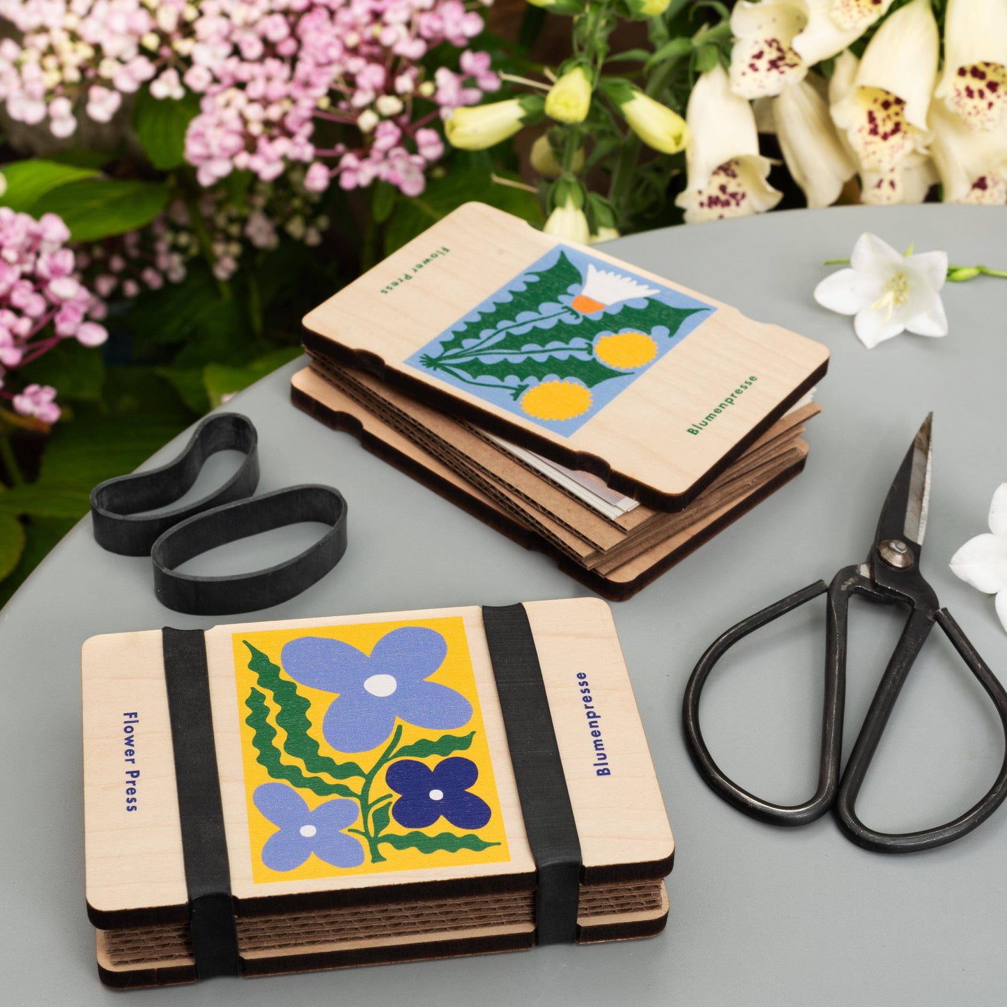 Two small books with floral designs, a rubber band, and scissors on a gray surface with flowers in the background.