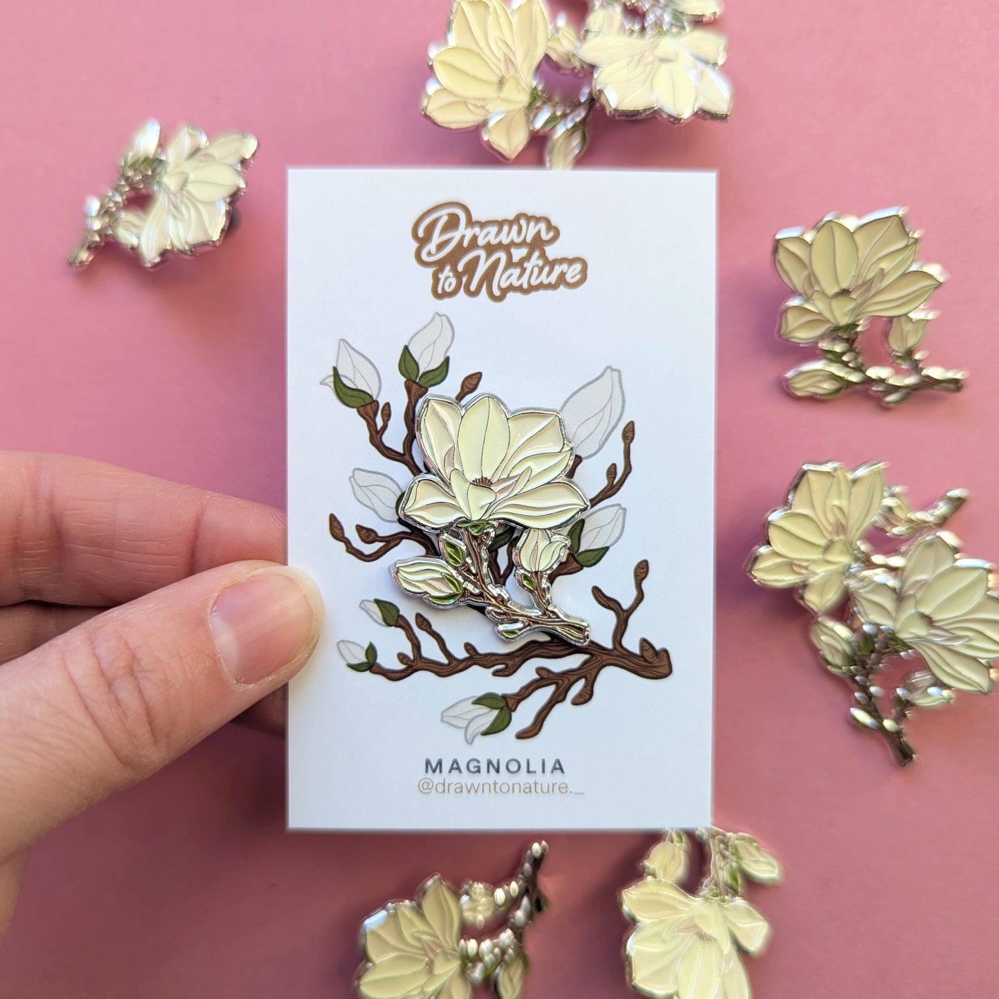 Gold magnolia flower pin on a card with 'Drawn to Nature' branding, against a pink background.