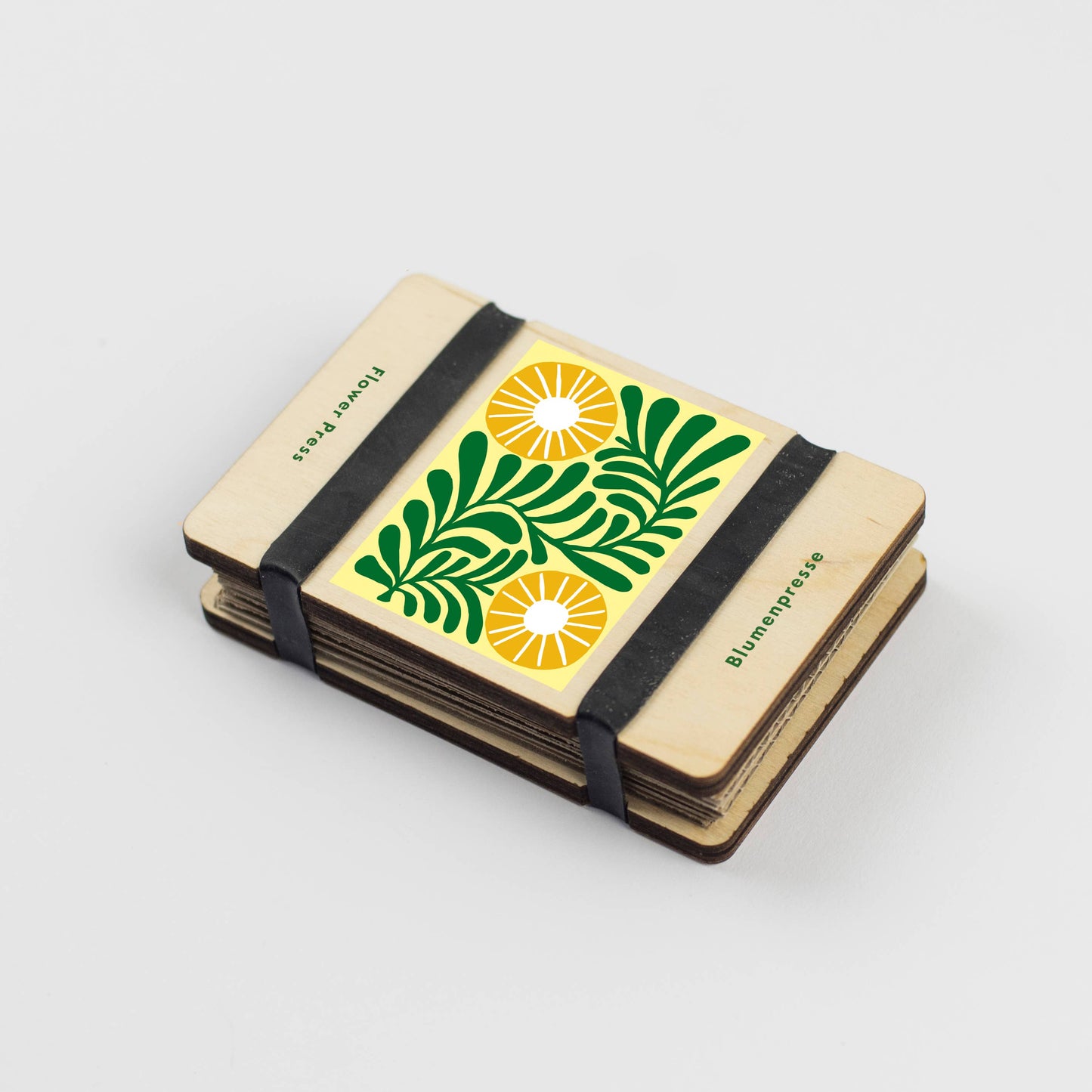 Small notebook with a decorative cover featuring green leaves and yellow suns, bound with black straps on a light gray background.