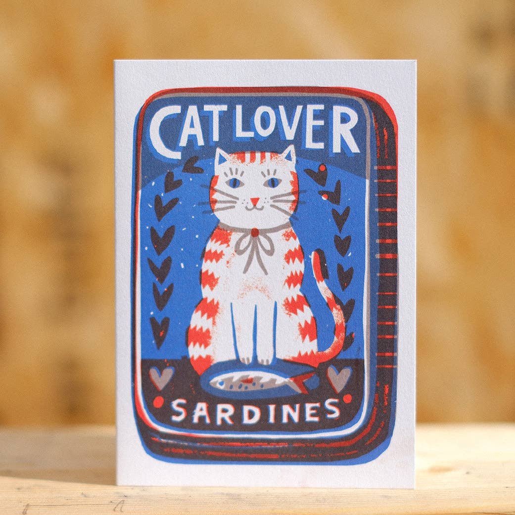 Card with a cat illustration and 'Cat Lover Sardines' text on a wooden surface.