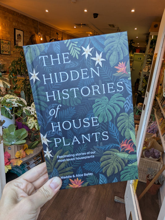 Book titled 'The Hidden Histories of House Plants' held in a hand with a store interior in the background.
