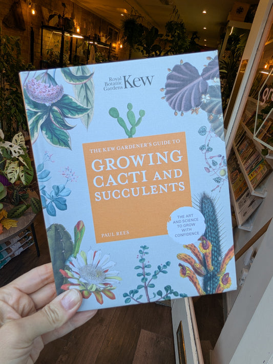 Book titled 'The New Gardener's Guide to Growing Cacti and Succulents' by Kew held in a hand with a plant store background.