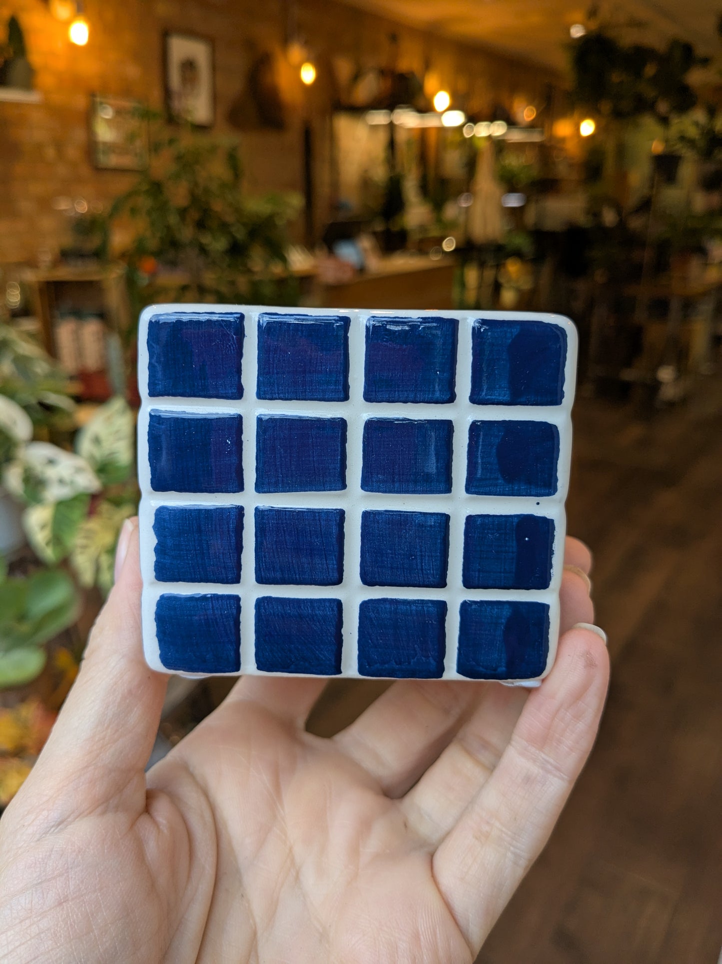 Hand holding a small ceramic tile with blue square patterns in a blurred indoor setting.