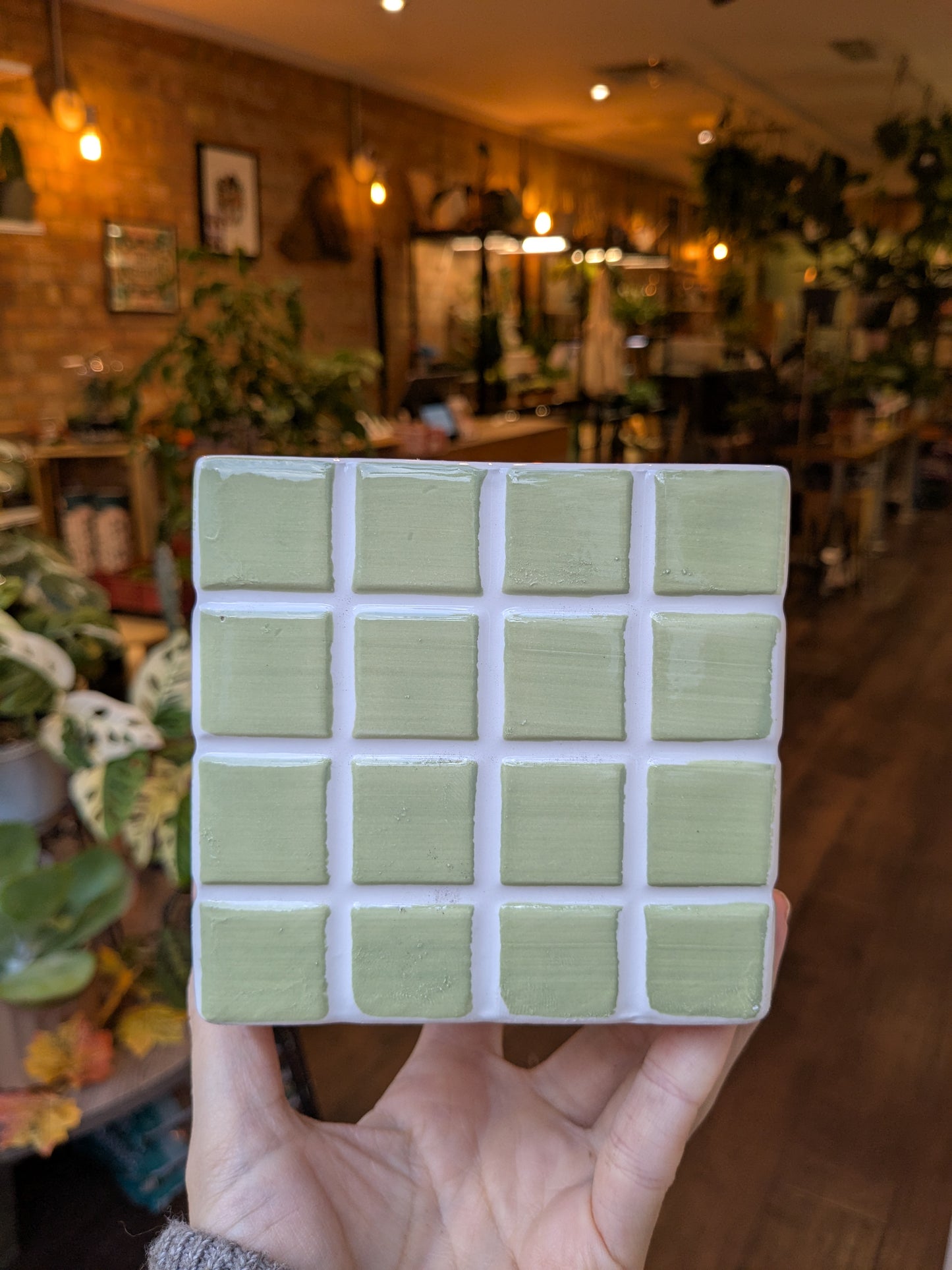 Hand holding a green ceramic tile with a blurred indoor setting in the background