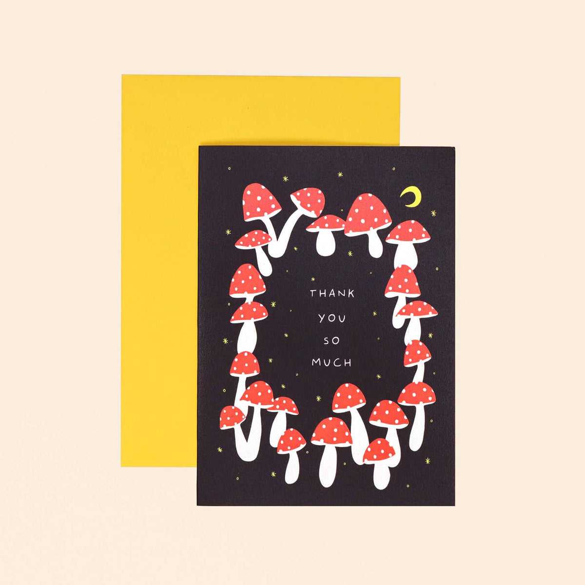 A greeting card with a black background, decorated with red and white mushrooms and the words 'THANK YOU SO MUCH' in white. A bright yellow envelope is placed behind the card on a light beige background.