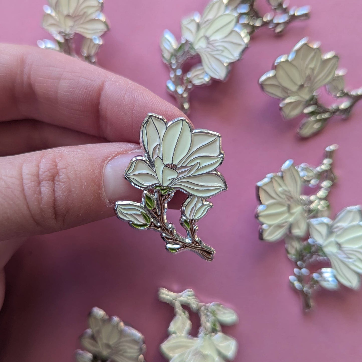 Floral enamel pin held between fingers with more pins on a pink background
