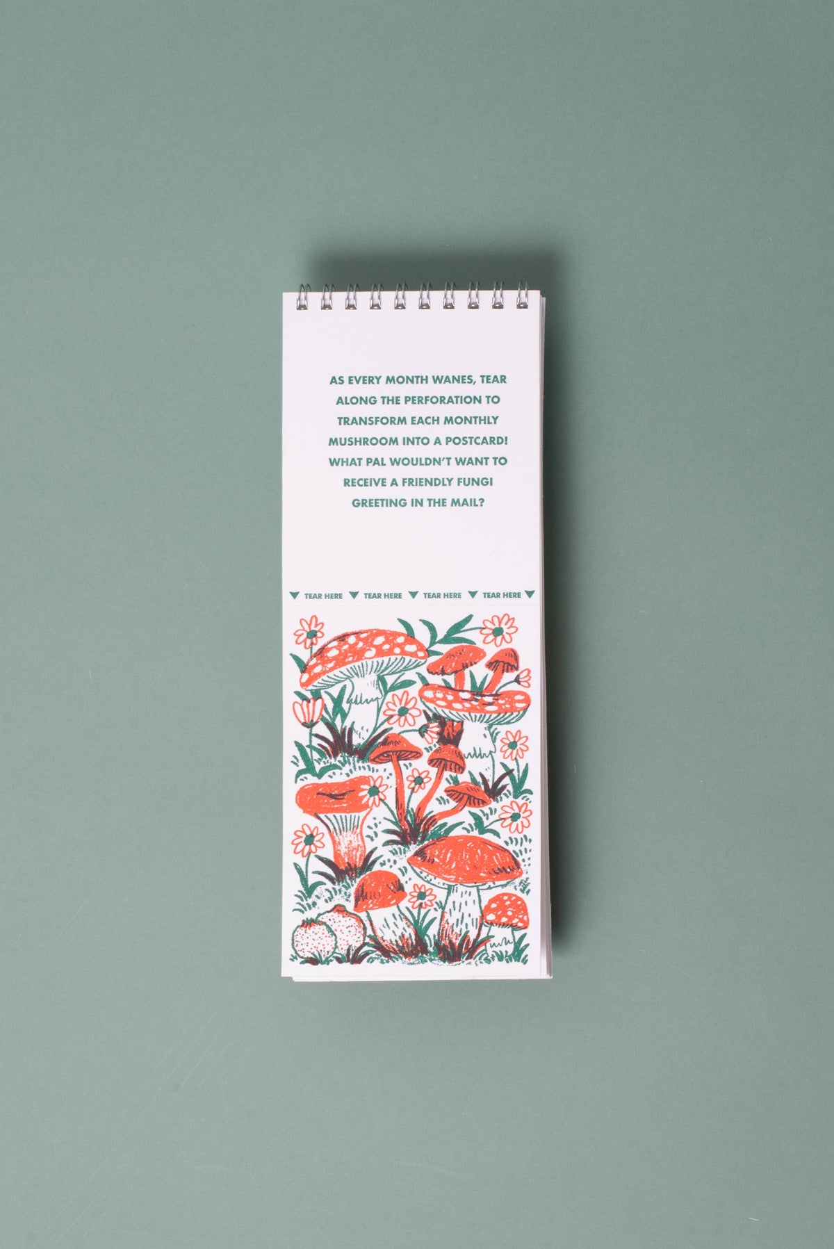 A tall, narrow spiral-bound notebook or desk calendar with a white cover, featuring a printed mushroom illustration in coral red, green, and black, and a short quote at the top, photographed on a muted sage green background.