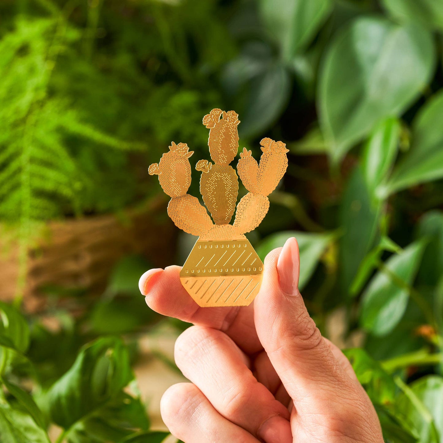 Gold cactus-shaped pin held by a hand with a green leafy background