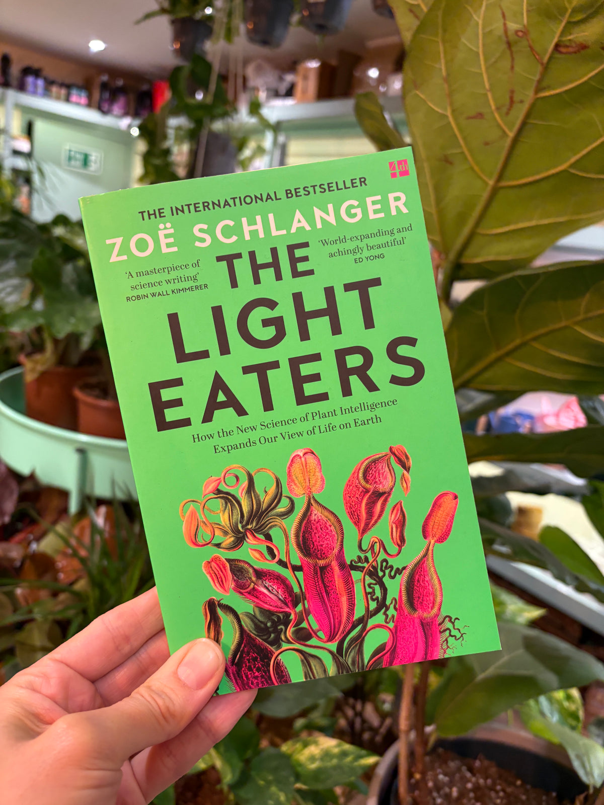 A hand holding a copy of the book 'The Light Eaters' by Zoë Schlanger, featuring a bright green cover with bold black title text and colorful botanical illustrations, set against a blurred background of indoor plants.