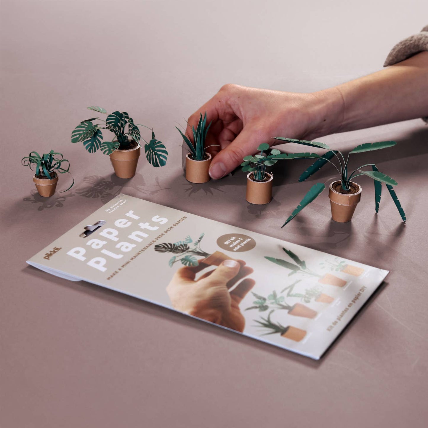 Hand interacting with small paper plants on a table next to an open book titled 'Paper Plants'.