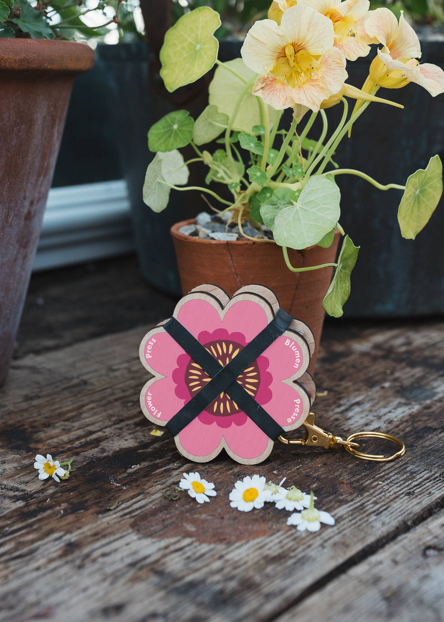 Floral keychain with a pink flower design on a wooden surface with potted plants in the background.