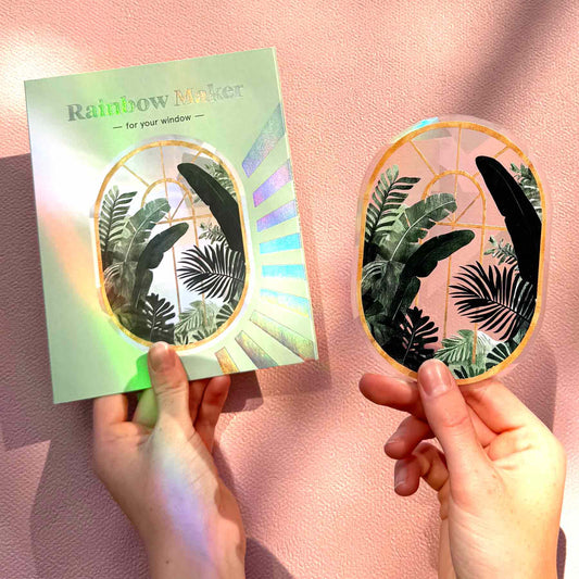 Hands holding a leaf-themed sticker with a colorful card labeled 'Rainbow Maker' on a pink background.