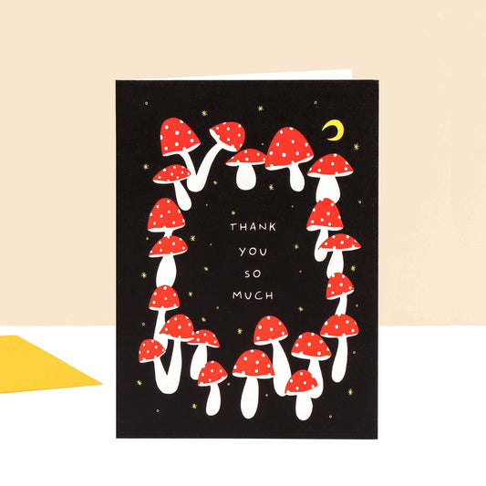 A black greeting card featuring a border of red and white mushrooms, small gold stars, a yellow crescent moon, and the text 'THANK YOU SO MUCH' in white. The card is shown standing upright against a light beige background with a small yellow envelope corner visible at the lower left.