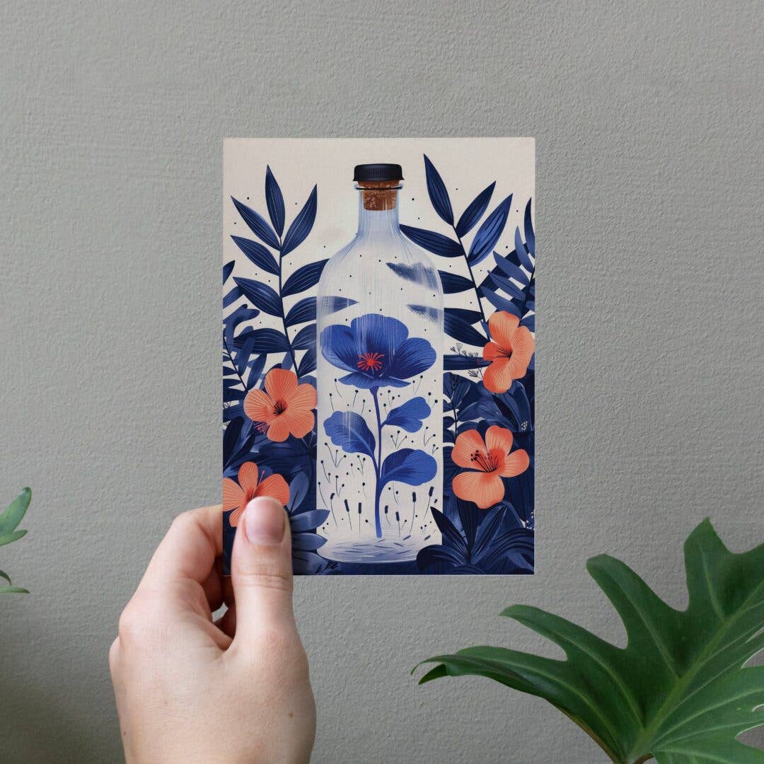 Hand holding a postcard with a floral design featuring a bottle and flowers against a gray wall.