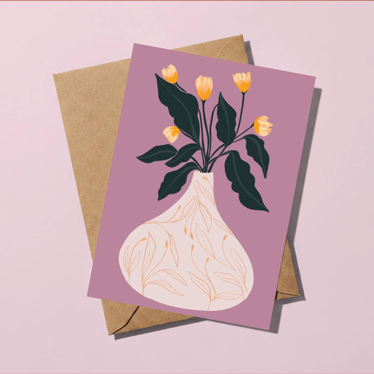 A square greeting card with an illustration of yellow tulips in a cream vase against a dusty purple background, shown with a brown kraft envelope on a light pink surface.