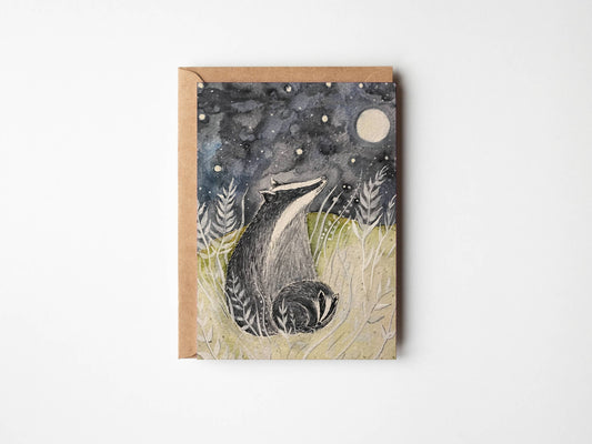 A square art print or card featuring a watercolor-style illustration of a badger in a grassy field under a starry night sky with a moon, shown with a kraft envelope on a white background.
