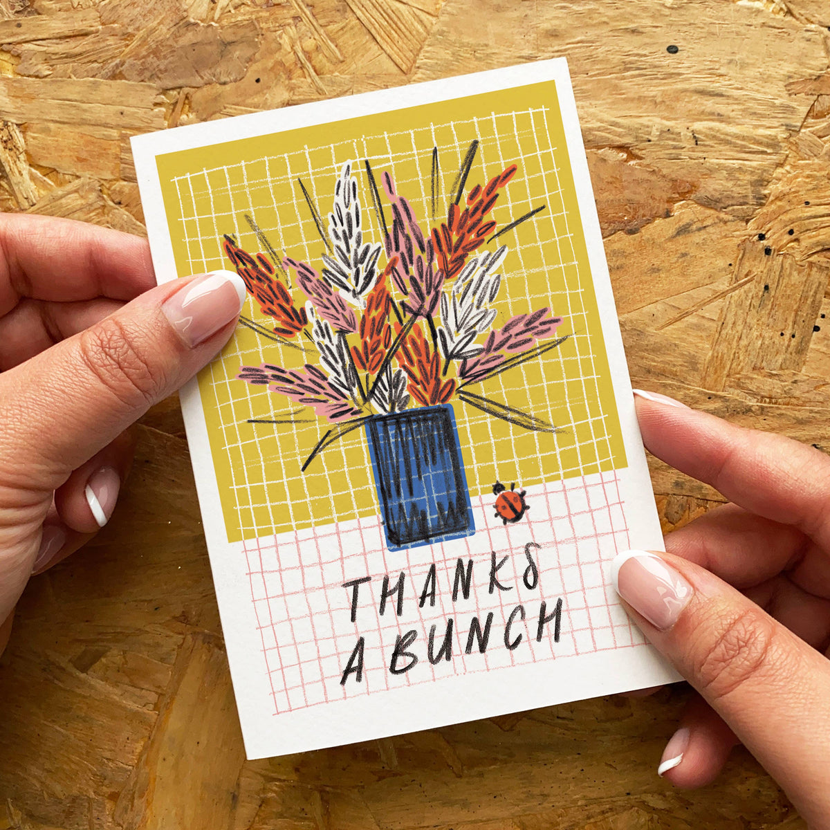 Hand holding a card with a plant illustration and 'Thanks a Bunch' text on a wooden surface