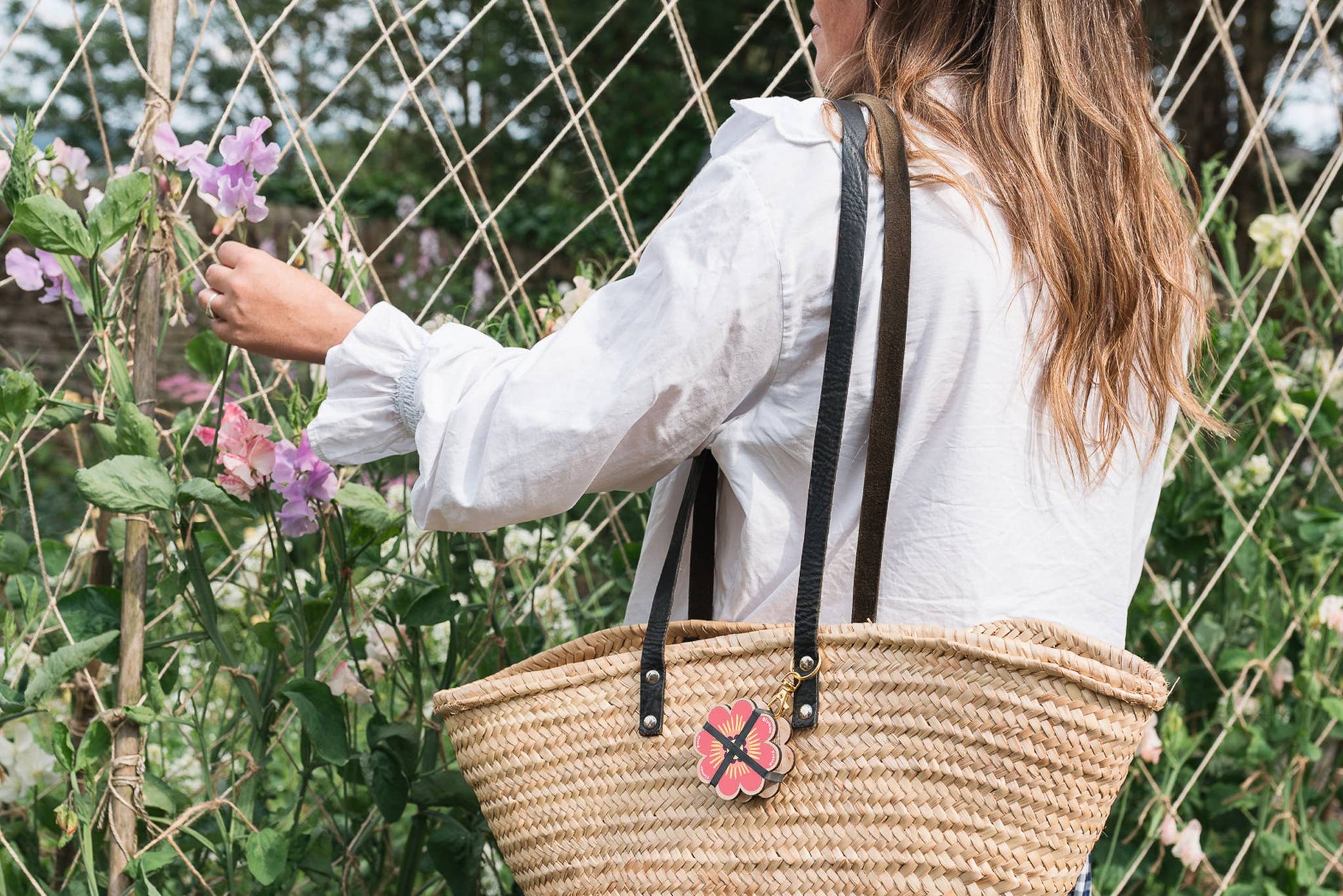 Person wearing a white jacket with a straw bag holding a pink flower charm, standing in front of a wire fence and plants.