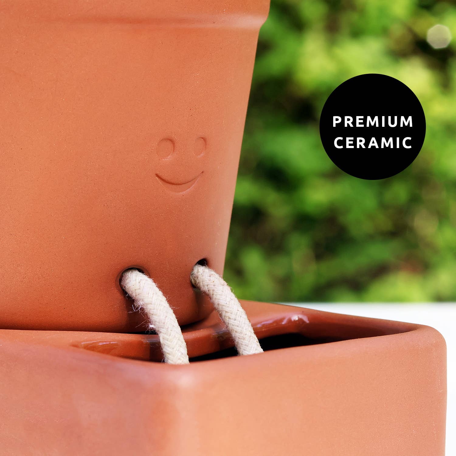Terracotta ceramic planter with smiley face design and text 'Premium Ceramic' on a blurred green background.