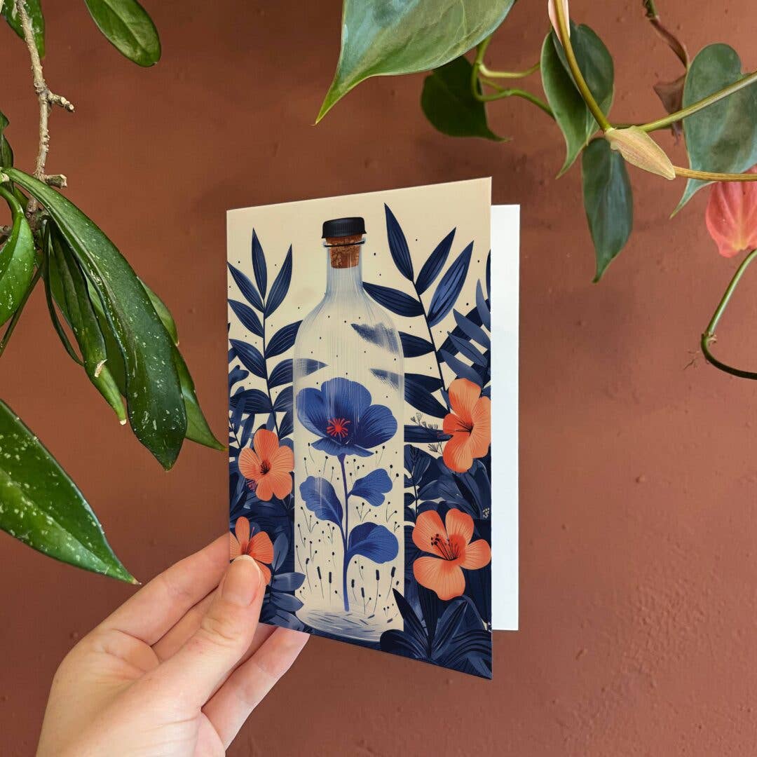 Hand holding a card with floral and bottle design against a brown background