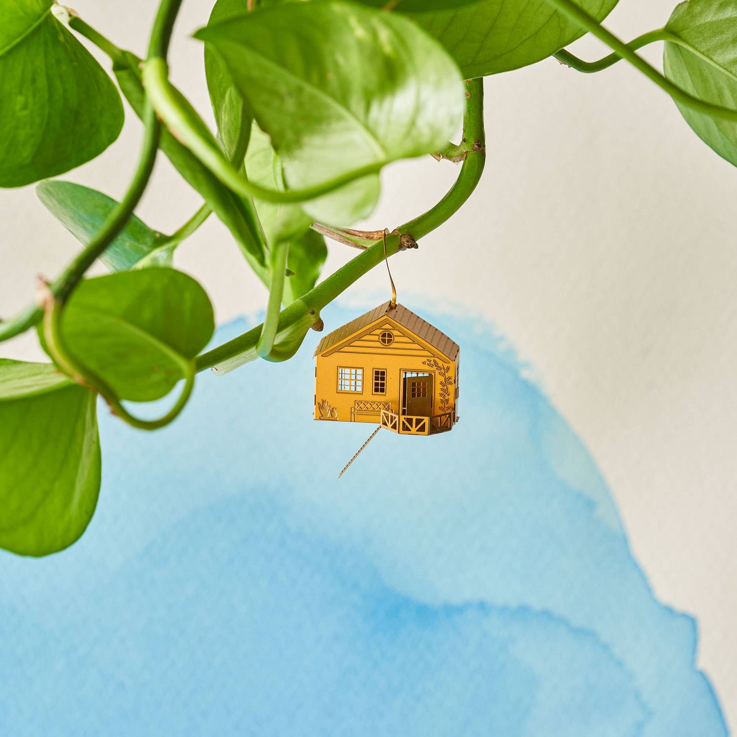 Small yellow house ornament hanging from a green leafy branch against a blue and beige background