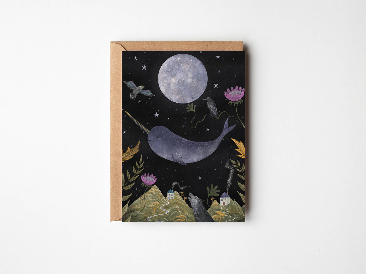 A square art print or card featuring a whimsical night scene with a large moon, a narwhal, stars, flowers, and small houses on hills, shown with a kraft envelope against a light gray background.
