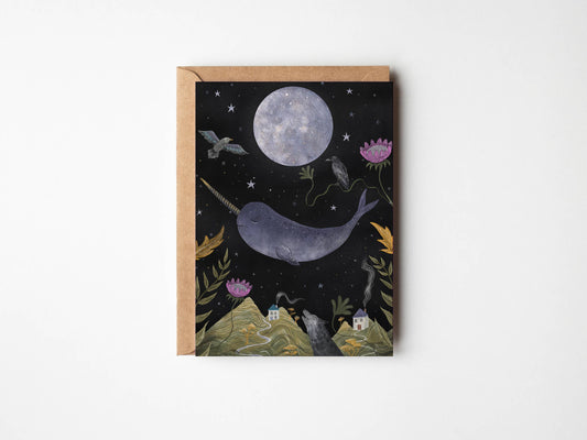 A square art print or card featuring a whimsical night scene with a large moon, a narwhal, stars, flowers, and small houses on hills, shown with a kraft envelope against a light gray background.
