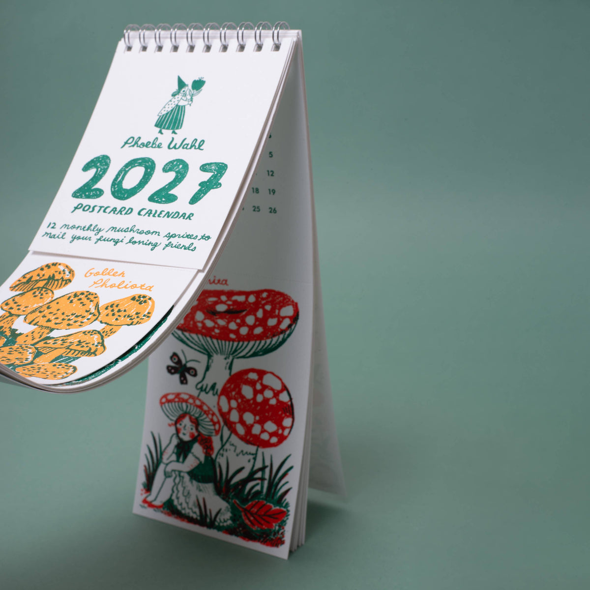 A spiral-bound 2027 postcard calendar by Phala Waltse stands open on a stand against a muted green background. The calendar pages feature whimsical illustrations, including mushrooms, flowers, a butterfly, and a fairy.