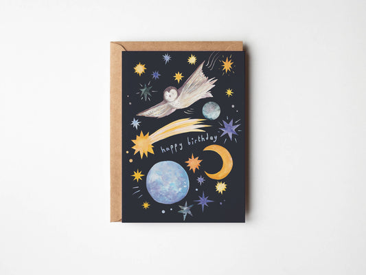 A square birthday card with a kraft paper envelope, featuring a dark navy space illustration with a shooting star, planets, stars, and a crescent moon, plus the words 'happy birthday' in white script.