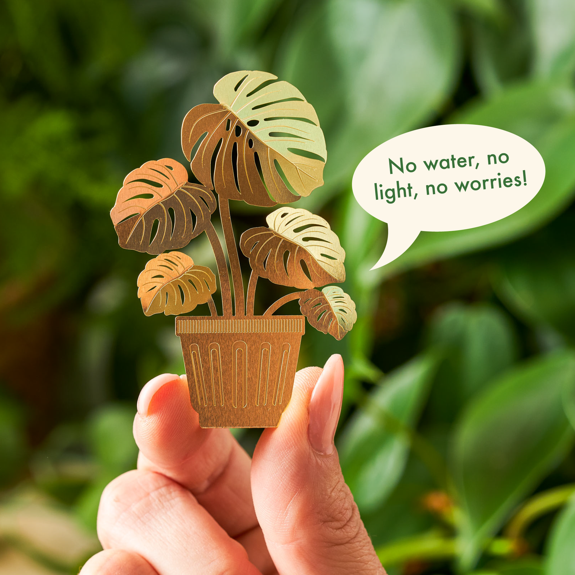 Hand holding a small potted plant figurine with a speech bubble saying 'No water, no light, no worries!' against a green leafy background.