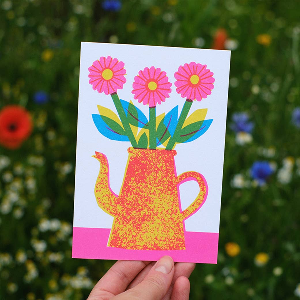 Colorful card with a teapot and flowers held against a natural background