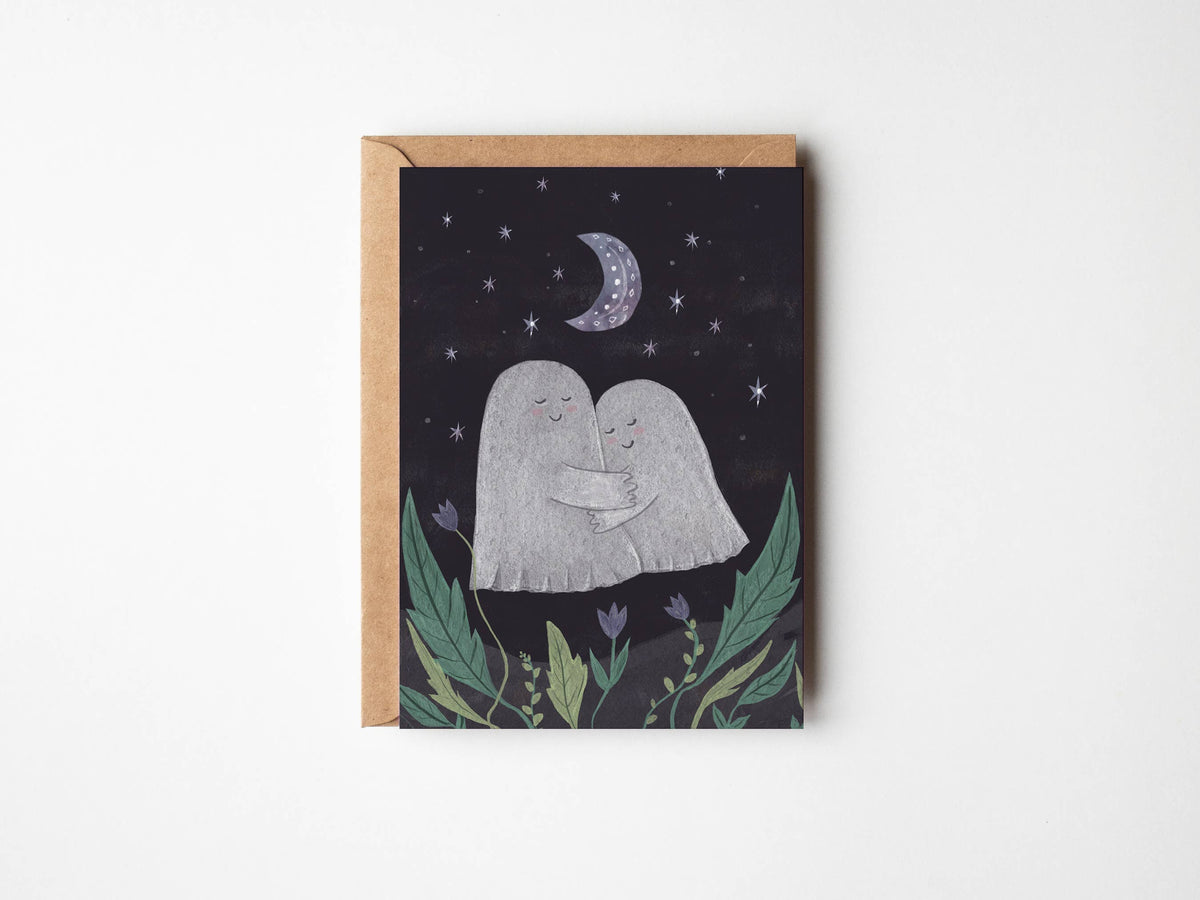 A square greeting card with a kraft paper envelope, featuring an illustration of two hugging ghosts under a crescent moon and stars, displayed on a clean white background.