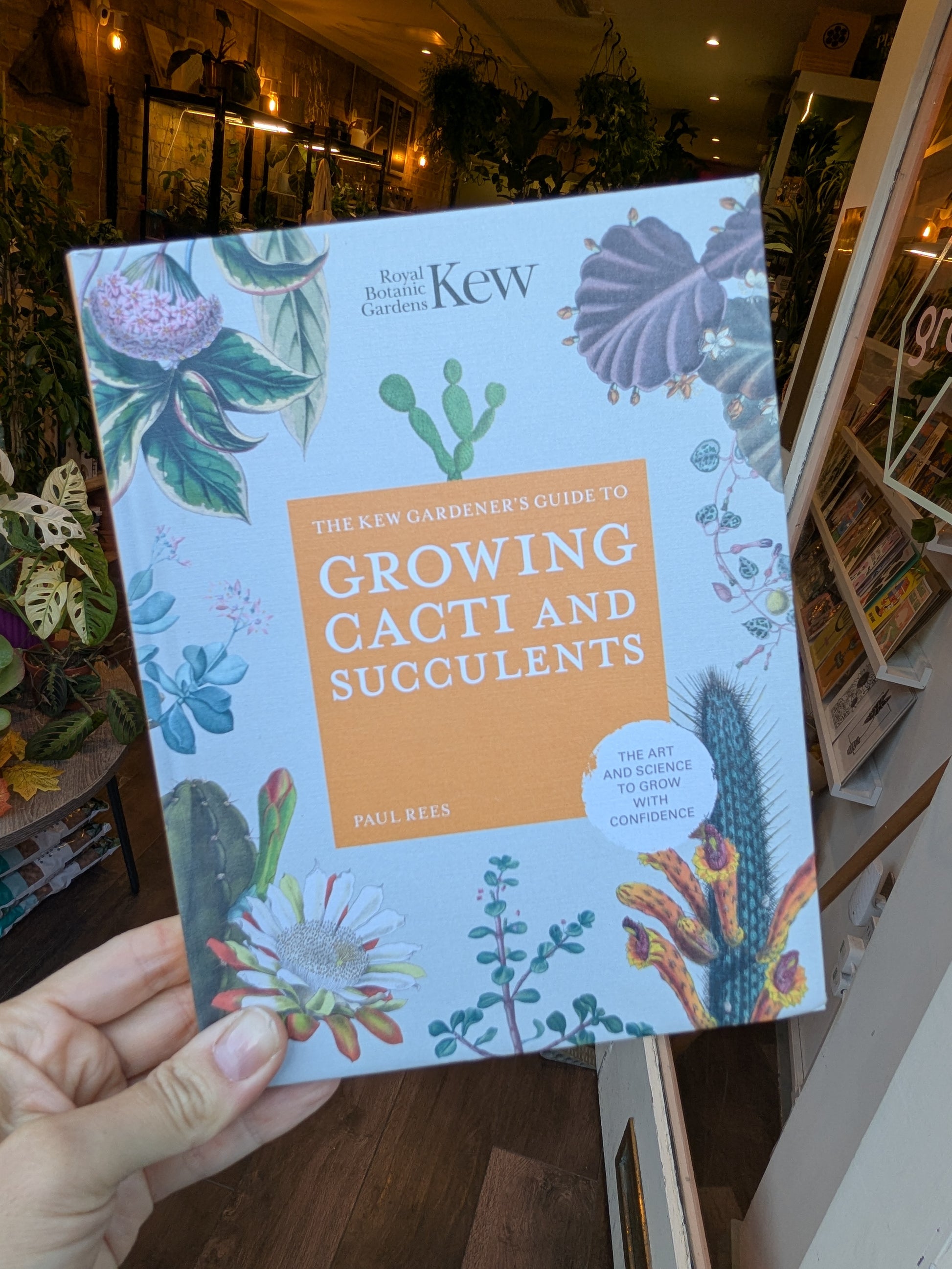 Book titled 'The New Gardener's Guide to Growing Cacti and Succulents' by Kew held in a hand with a plant store background.