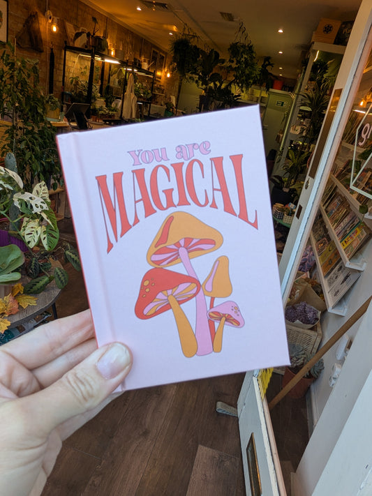 Hand holding a book with 'You are Magical' text and mushroom illustration in a store setting