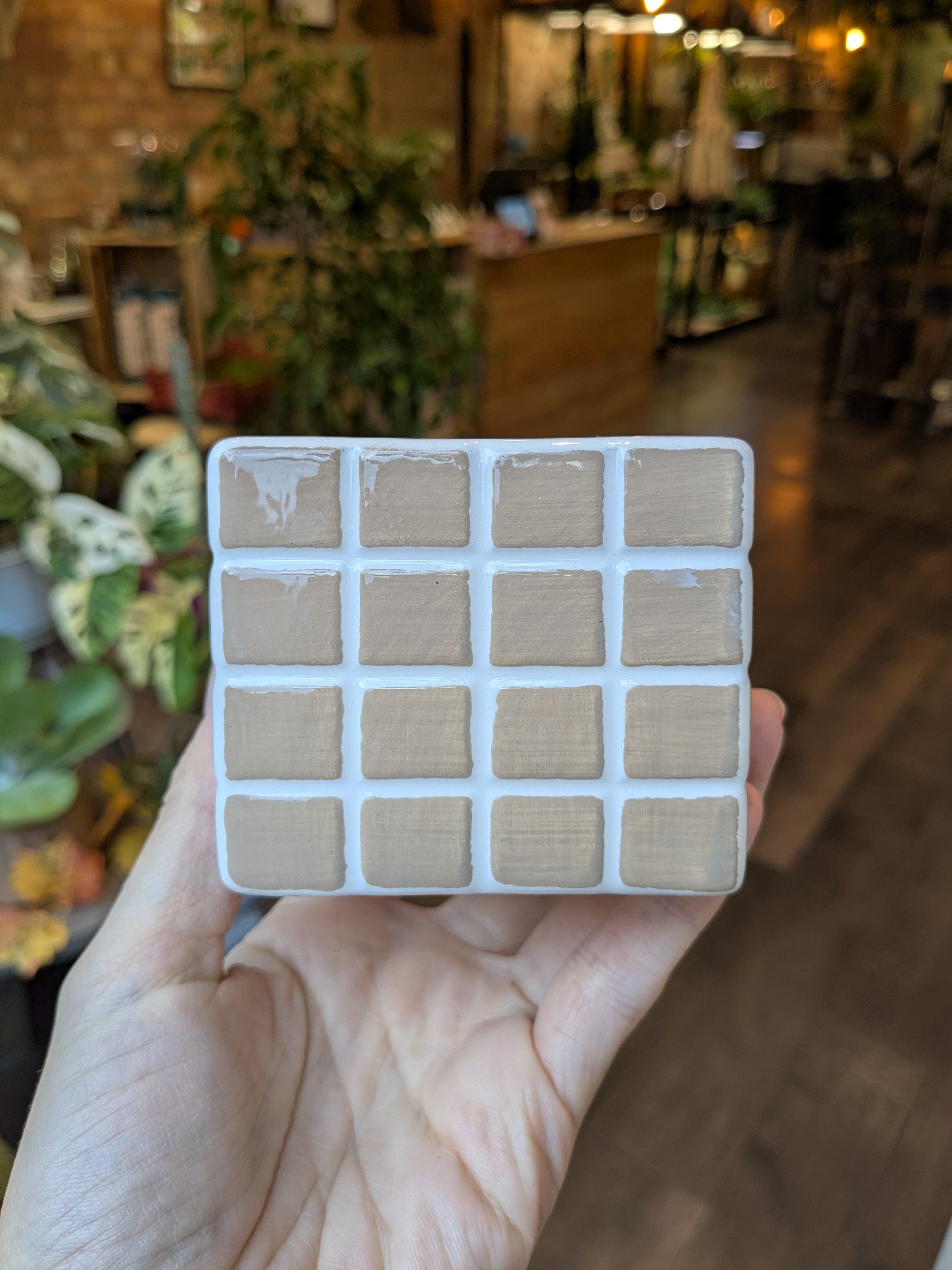 Hand holding a small square tile with a neutral color swatch pattern, blurred indoor background