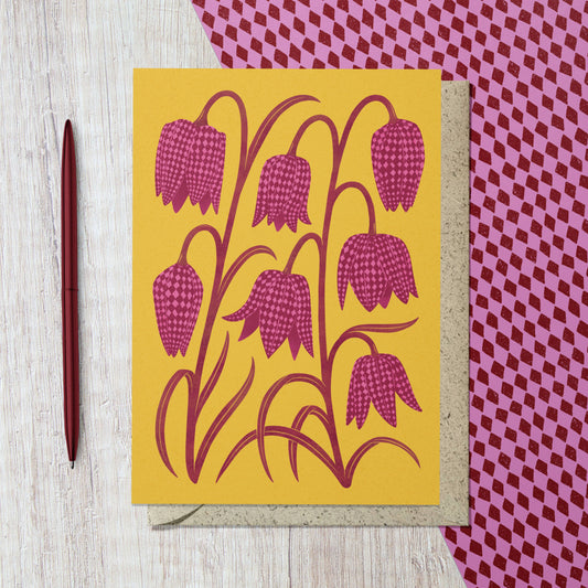 Card with pink floral design on a yellow background, placed on a wooden surface with a pen and checkered fabric.