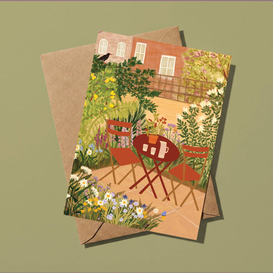A rectangular art print or card featuring an illustrated garden patio scene with a red folding table, chairs, flowers, and a brick building, shown overlapping a brown kraft envelope on a muted olive background.