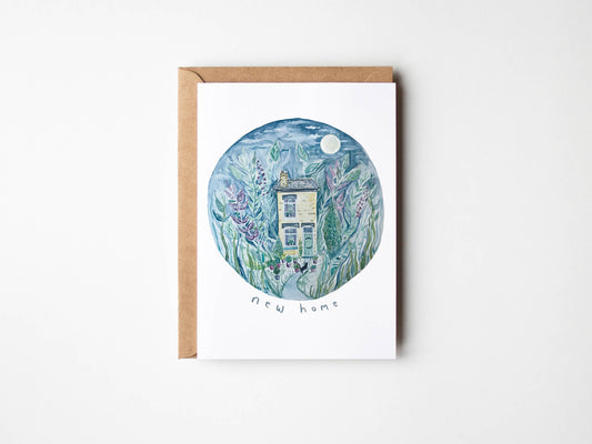 A white greeting card with a brown kraft envelope, featuring a circular watercolor illustration of a small house surrounded by green and purple plants, with the words 'new home' printed below.