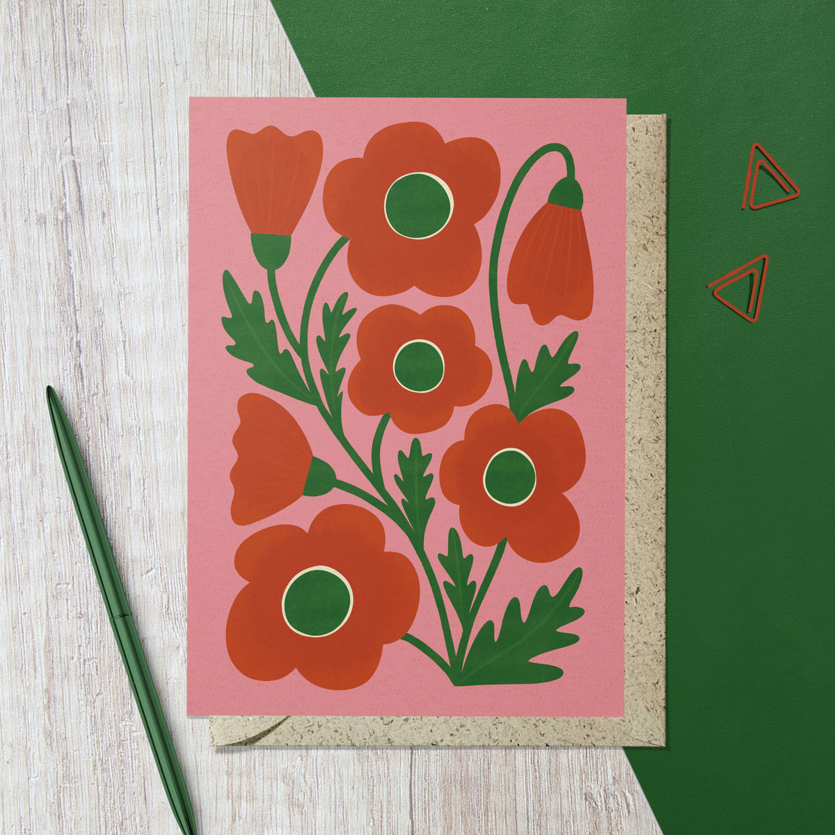 Card with red floral design on pink background, green envelope, and pen on wooden surface