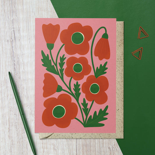 Card with red floral design on pink background, green envelope, and pen on wooden surface