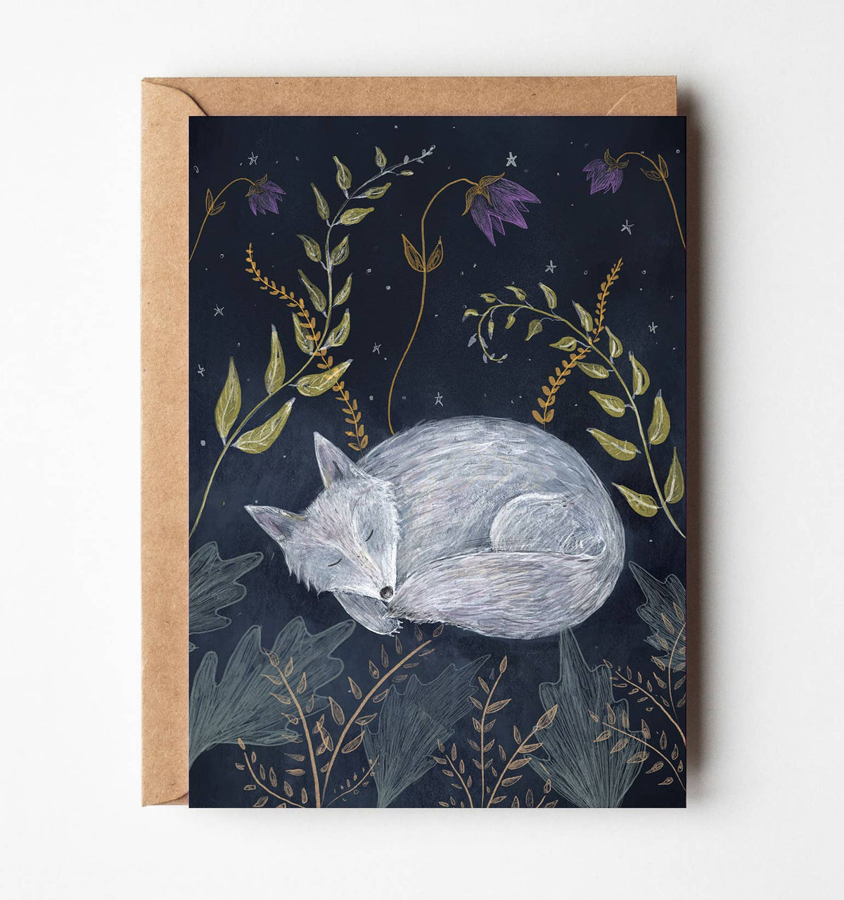 A square art print or card featuring a sleeping gray fox on a dark navy night scene with purple flowers and botanical details, shown with a kraft envelope against a light neutral background.