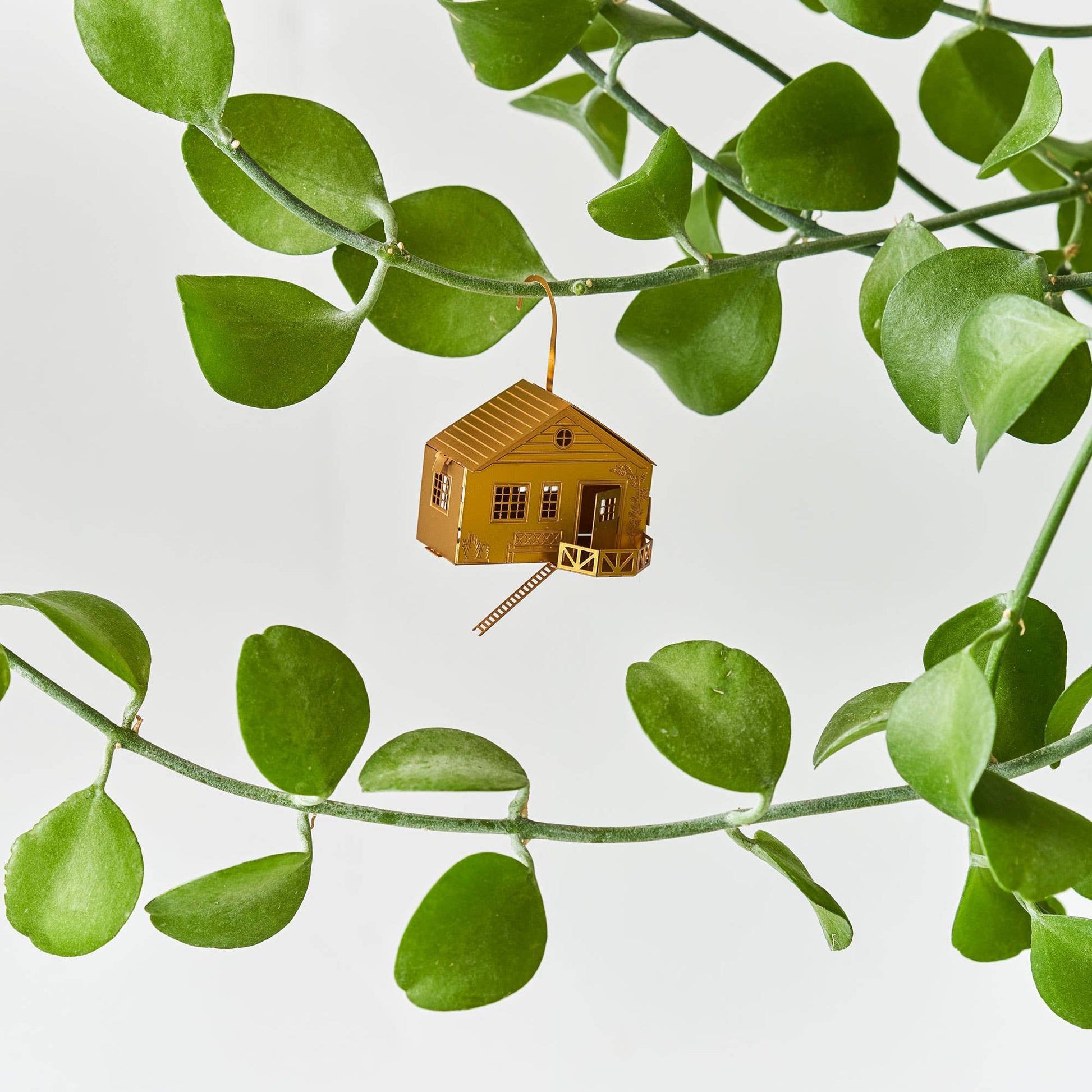 Small wooden cabin ornament hanging among green leaves on a light gray background