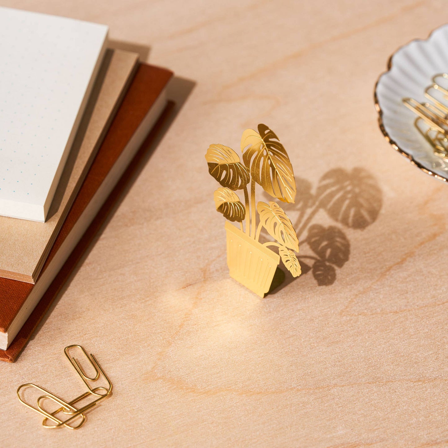 Gold leaf-shaped paper clip on a beige surface with a notebook and decorative item.