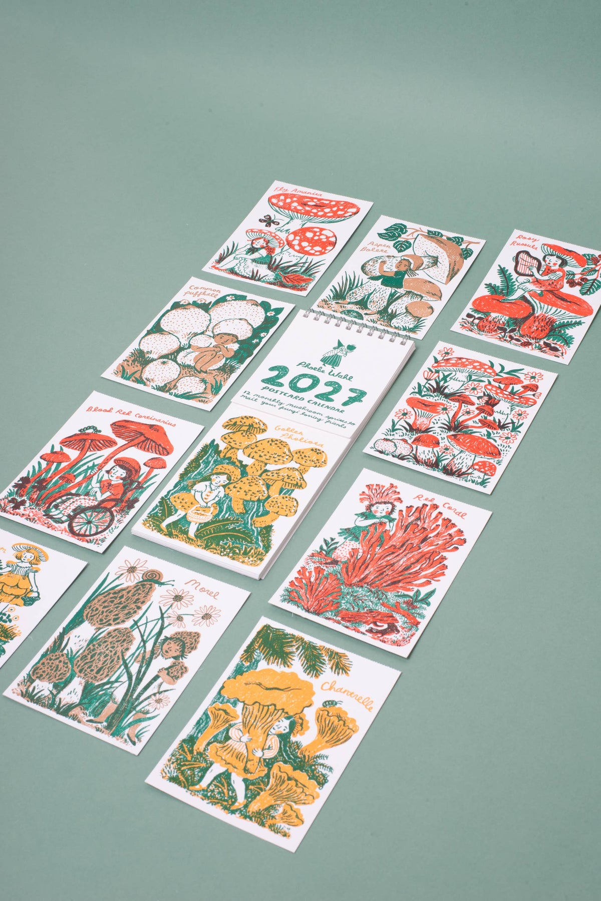 A set of illustrated cards or prints arranged on a muted green background. The cards feature colorful drawings of mushrooms, flowers, and nature scenes, with one central card showing the year 2023.
